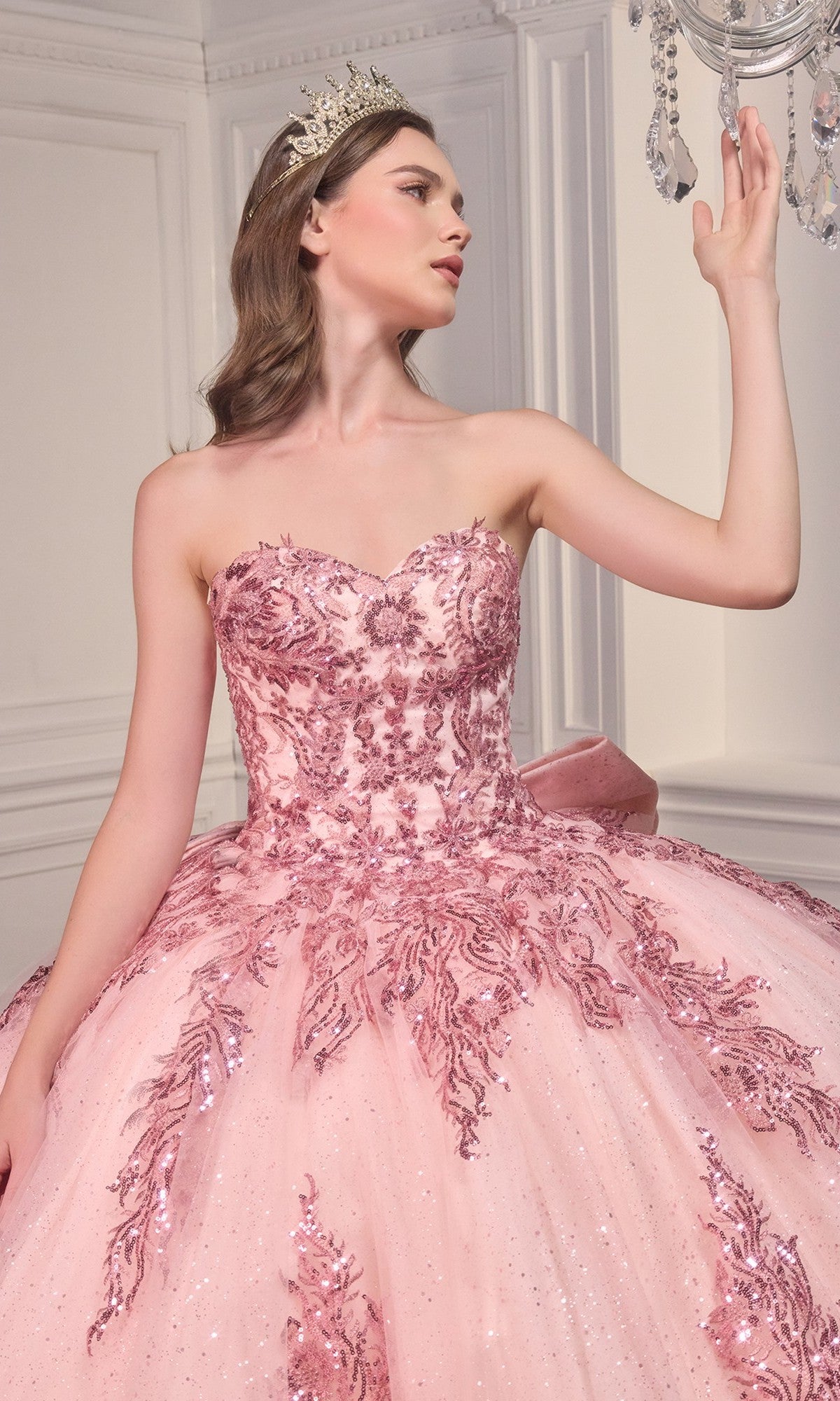 A woman in the Ladivine Strapless Quinceanera Dress 15715, a pink ball gown with sparkling sequins and a tiara, stands indoors and gracefully touches a crystal chandelier.